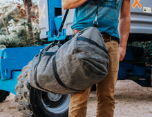 Person carrying a large, rugged gray duffel bag with black straps and a shoulder strap, wearing a blue shirt and beige pants, standing next to industrial equipment