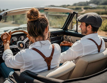 Man and women wearing brown leather suspenders, vintage outfit in convertible car, scenic outdoor setting