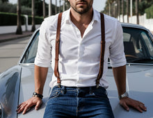 Man wearing brown leather suspenders, white shirt and jeans, leaning against a car