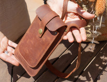 Alt Text: Close-up of hands holding a handmade leather phone crossbody bag. Description: A close-up view of a person's hands holding a handmade leather phone crossbody bag. The bag is brown with a front flap secured by a brass button and a long strap. The background includes part of a wooden table and some decorative flowers.