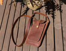 Alt Text: Handmade leather phone crossbody bag on a wooden table. Description: A handmade leather phone crossbody bag lying on a wooden table. The bag is brown with a front flap secured by a brass button and a long strap. The background includes shadows and part of a decorative basket.