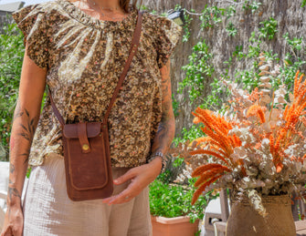 Alt Text: Woman wearing a handmade leather phone crossbody bag. Description: A woman wearing a floral blouse and light-colored pants is showcasing a handmade leather phone crossbody bag. The bag is brown with a front flap secured by a brass button and features a long strap worn across the body. The background includes greenery and orange flowers.