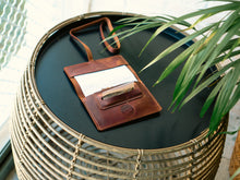 Handmade brown leather tobacco pouch resting on a round black tabletop, open to show rolling paper and tobacco, with a long leather strap