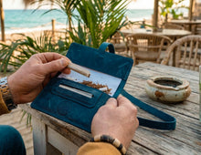 Open blue leather tobacco pouch held over a rustic beachside table, showing loose rolling tobacco, rolling papers, a rolled cigarette, and a built-in paper holder, with a long leather strap,