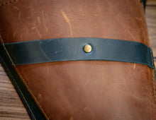 Close-up of a brown leather tool pouch item with a dark navy leather strap across the middle secured by a brass snap button, set against a wooden surface.