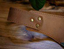 A close-up of a brown leather tool belt with brass rivets and stitching, resting on a rustic wooden surface.