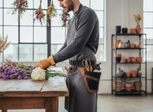brown leather tool pouch stands at a wooden table, handling purple and white flowers in a bright, modern greenhouse or workshop.