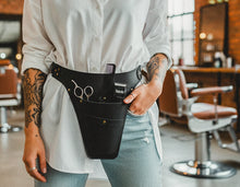 A black leather hair stylist's waist belt with multiple pockets holding scissors, a comb, and other tools, worn by a person in a salon setting.