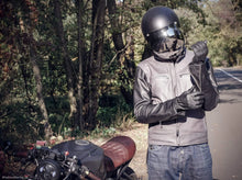 Person wearing a helmet and motorcycle jacket standing next to a motorcycle on a road with trees in the background