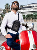 Man wearing a white shirt and blue jeans, with a black leather crossbody bag featuring detailed stitching and multiple pockets, sitting on a red and white scooter