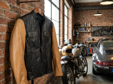 A black leather motorcycle vest with tan leather sleeves hangs on a hanger against a brick wall in a garage setting, with a motorcycle and a classic car in the background.