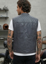 Man wearing a gray leather vest standing next to a motorcycle indoors.