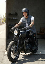 Man sitting on a motorcycle wearing leather biker gray vest a helmet and casual clothing.