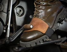 A close-up of a brown leather motorcycle boot with a tan brown leather  left boot cover and a black heel strap, shown resting on a motorcycle footpeg.