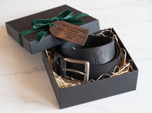 A black leather belt with a silver buckle and an embossed cross design, presented in an open black gift box 