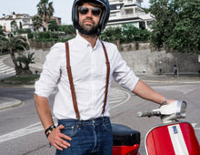 Man wearing a white shirt, blue jeans, and brown leather suspenders, standing next to a red and white scooter while wearing sunglasses and a helmet.