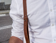 Close-up of a person wearing a white shirt, highlighting the brown leather strap of a crossbody bag with multiple djustment holes and a metal rivet