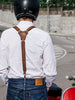 The image shows the back view of a person wearing brown black Reinforced leather suspenders over a white shirt, paired with blue jeans. They are sitting on a red and white scooter, with a helmet on, ready to ride. The suspenders intersect at a leather patch in the center of the back, which is embossed with a logo 