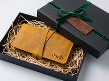 Personalized mustard-yellow leather tobacco pouch presented in an open black gift box with shredded paper filler, finished with a green velvet ribbon and a brown gift tag